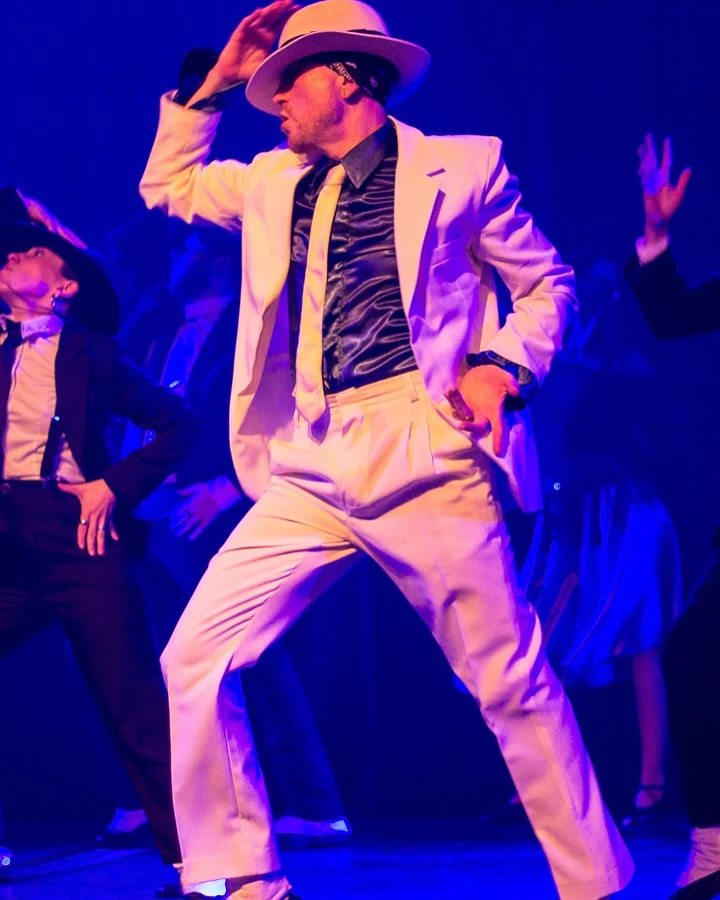 Intérprete con traje blanco y sombrero baila junto al elenco bajo luces azules
